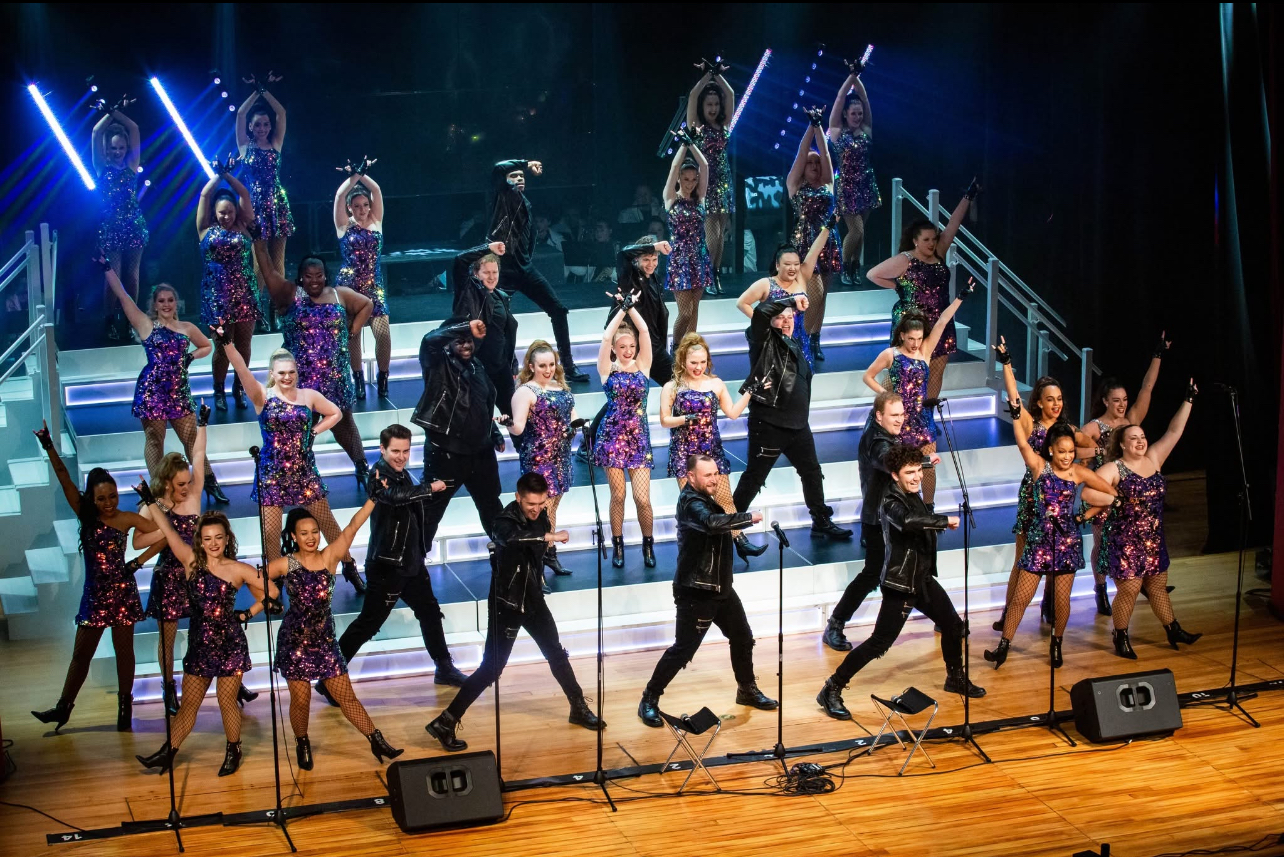 A group of performers in coordinated costumes dance energetically on a brightly lit, tiered stage.