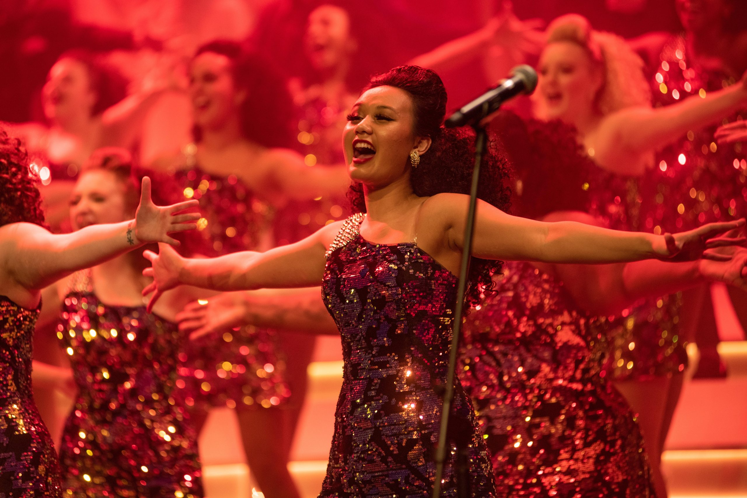 A group of performers in sequined outfits sing and dance energetically on stage under red lighting, with one performer at the front extending their arms.
