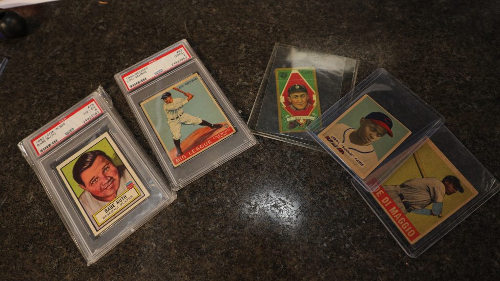 Five baseball cards are laid out on a counter in protective coverings. They feature Babe Ruth, Lou Gehrig, Ty Cobb, Satchell Paige, and Joe DiMaggio.