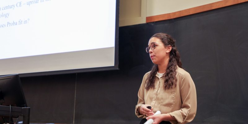 A person stands in front of a chalkboard and projection screen, holding papers and giving a presentation in a classroom.