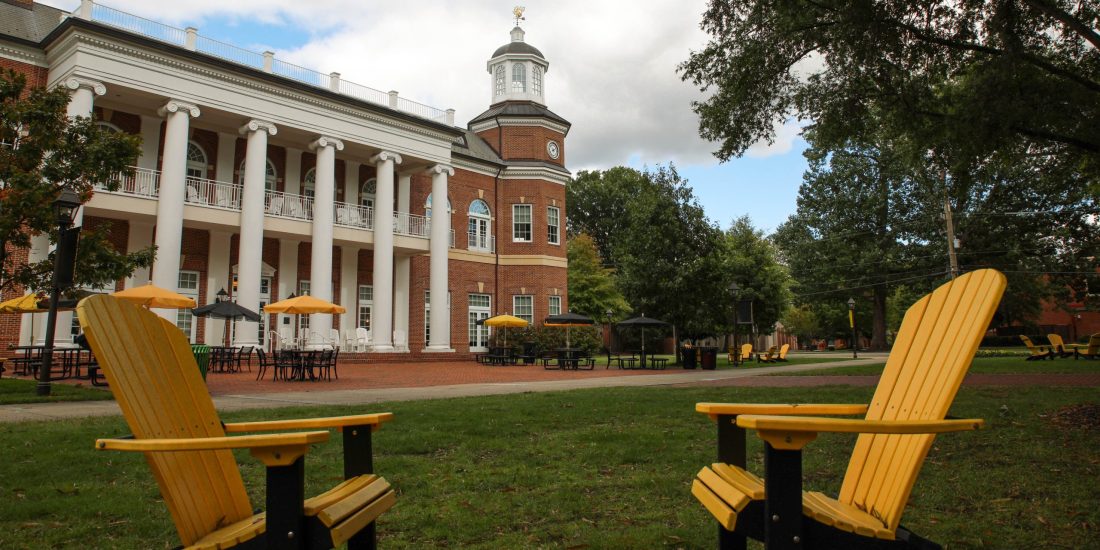 Yellow Adirondack chairs on a lawn face a red-brick building with columns and a clock tower, surrounded by trees and seating.
