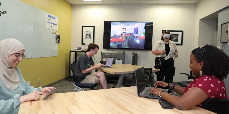Four people in a meeting room use devices; one stands with a VR headset before a screen showing a virtual environment.