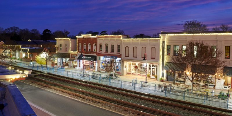 Storefronts illuminated at dusk line a small-town main street, with railroad tracks in front and a purple sky above.