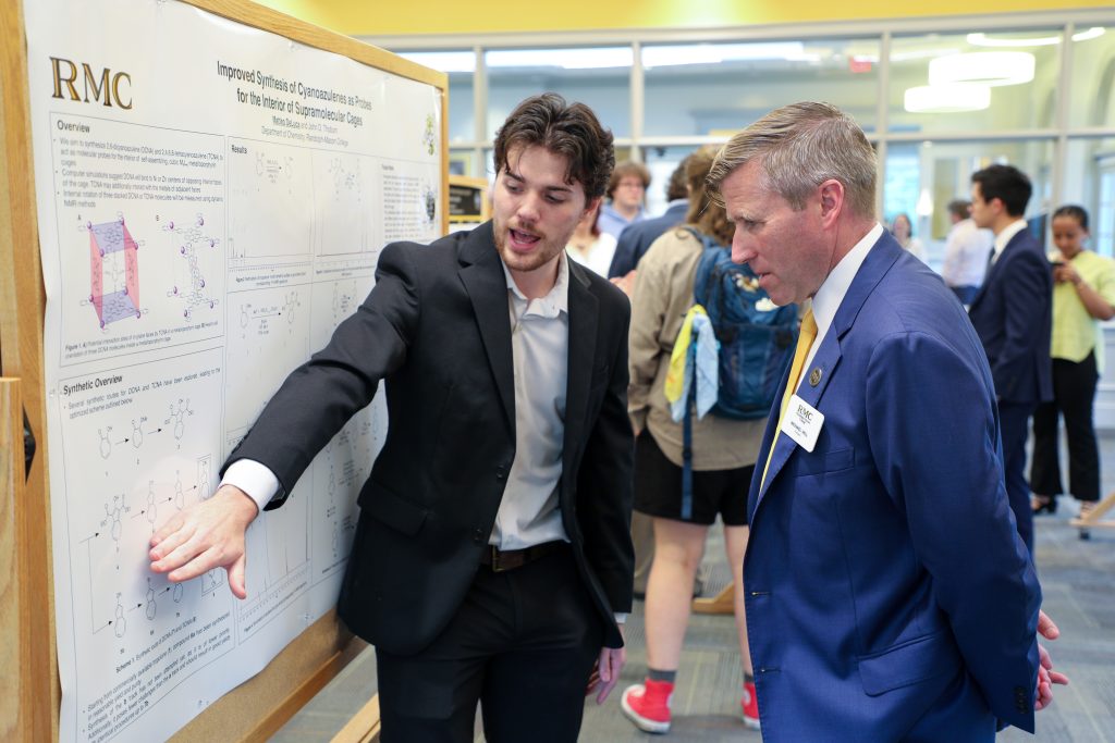 Two individuals in suits discuss a scientific poster at an indoor academic event, with other attendees in the background.