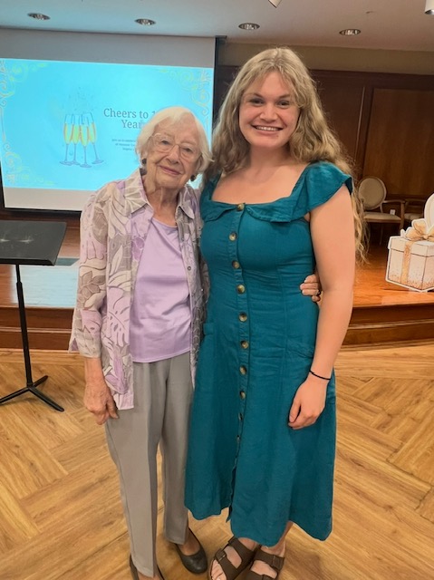 Two people stand indoors, smiling at the camera with a presentation slide reading "Cheers to Years" visible in the background.