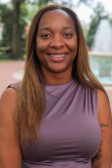 Person smiling at the camera outdoors, standing near a fountain with trees in the background.