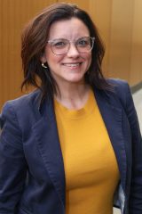 An individual wearing glasses and a navy blazer over a yellow top stands indoors, smiling at the camera.