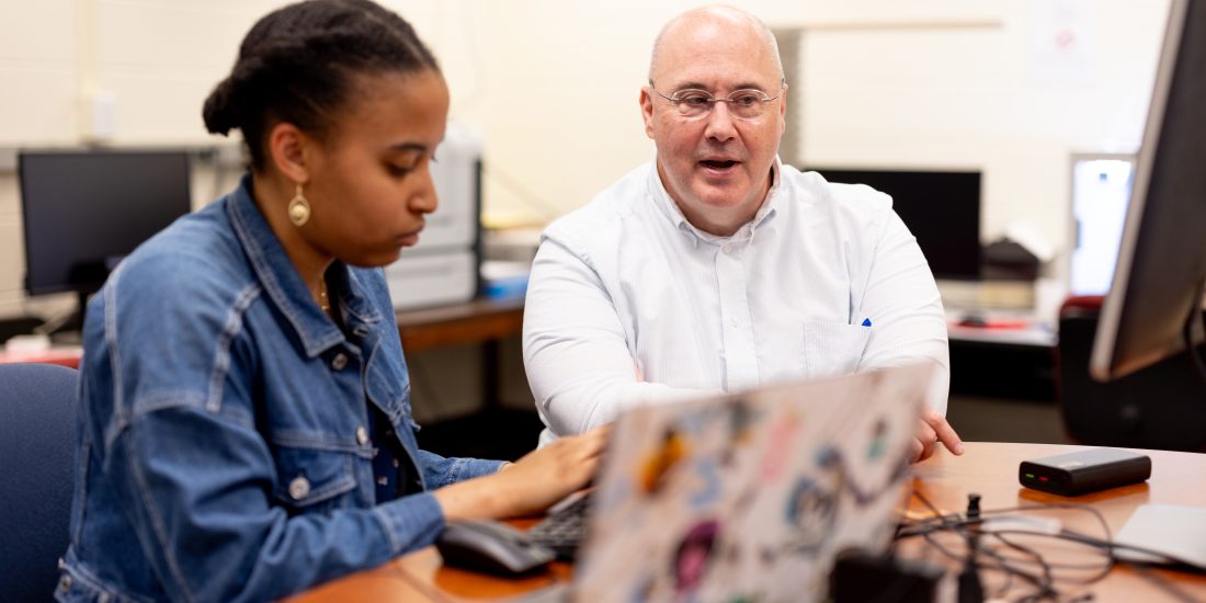 Two people work together at a laptop in a computer lab, one typing while the other speaks.