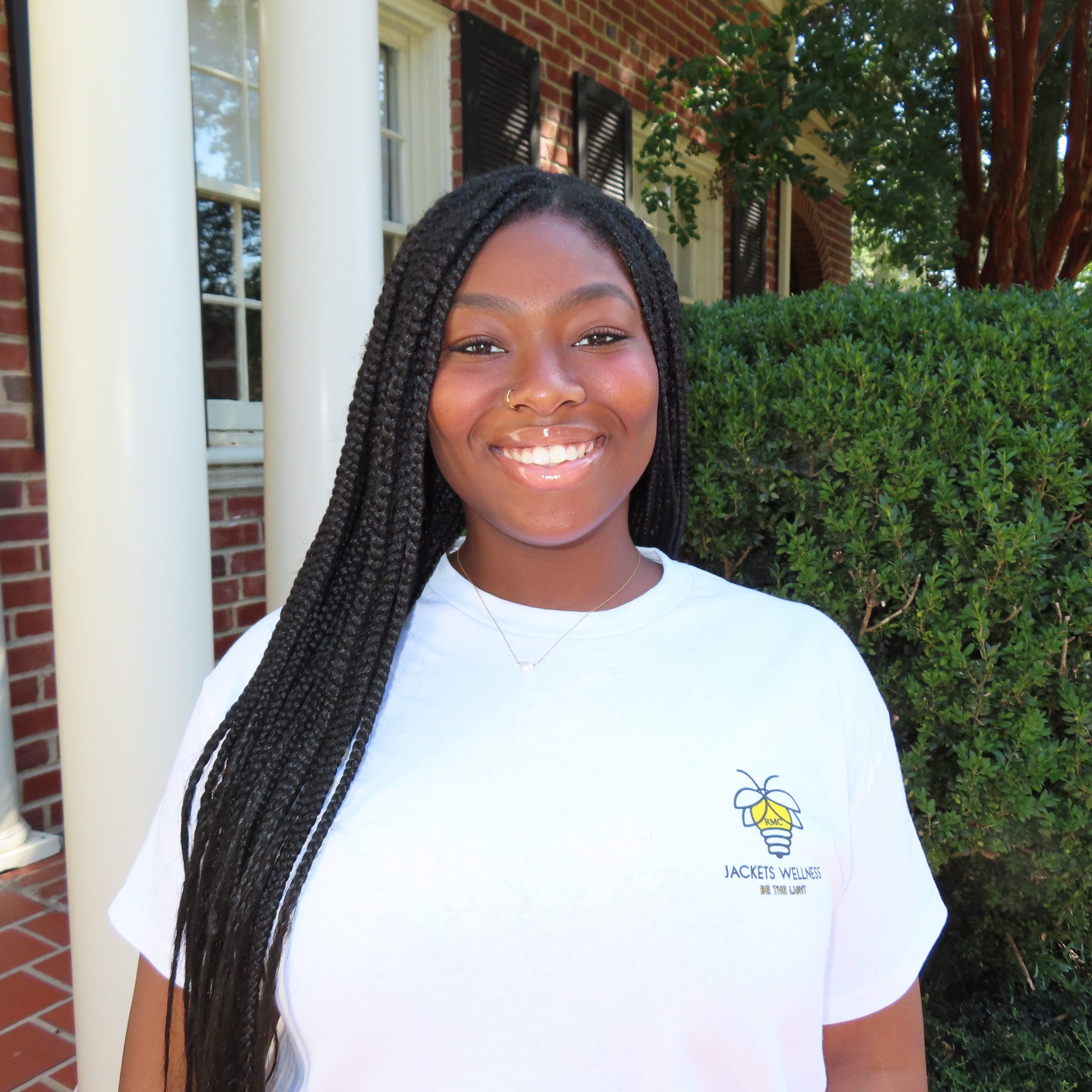 Person smiling outside in front of a brick building and greenery, wearing a white T-shirt with a small logo.