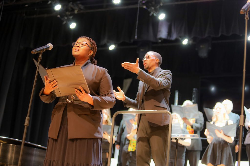 A vocalist sings into a microphone holding sheet music as a conductor leads a choir in the background on stage.