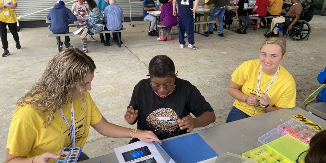A group, including RMC Nursing students and SOAR365 participants, participates in outdoor arts and crafts activities at tables, including painting and working with beads and supplies.