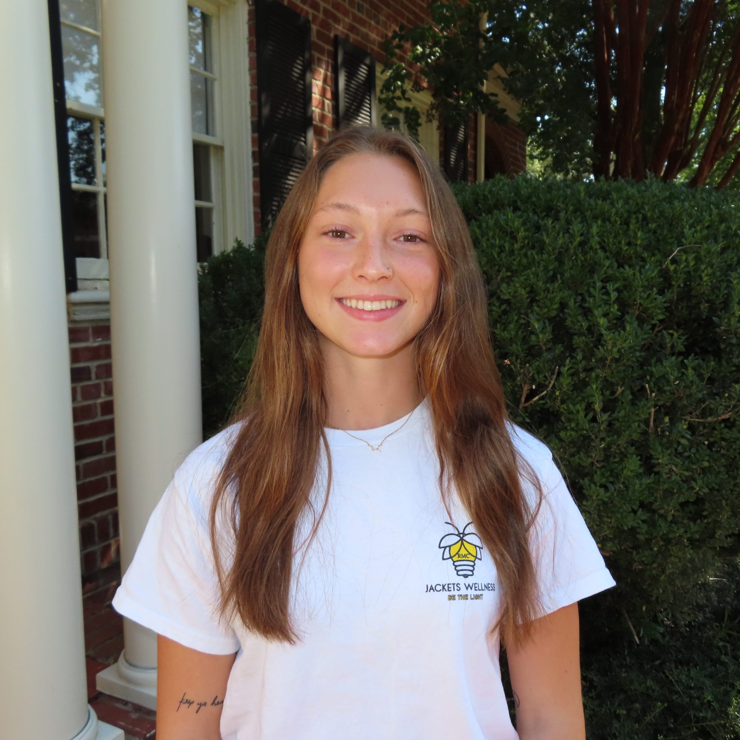 Person wearing a "Jackets Wellness" t-shirt stands outside in front of bushes and a brick building, smiling at the camera.