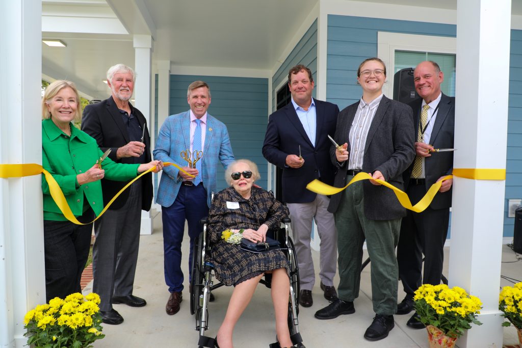 A group participates in a ribbon-cutting ceremony in front of a blue building, holding scissors and a yellow ribbon.