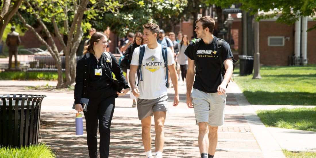 College students walk and talk on a sunny campus sidewalk with trees, grass, and other people in the background.