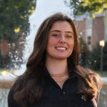 A student smiles outdoors in front of a fountain, with trees and a building in the background.