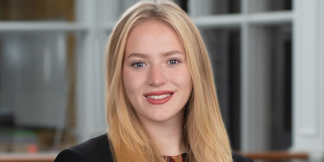 An individual in business attire stands indoors before blurred windows and railings, smiling at the camera.