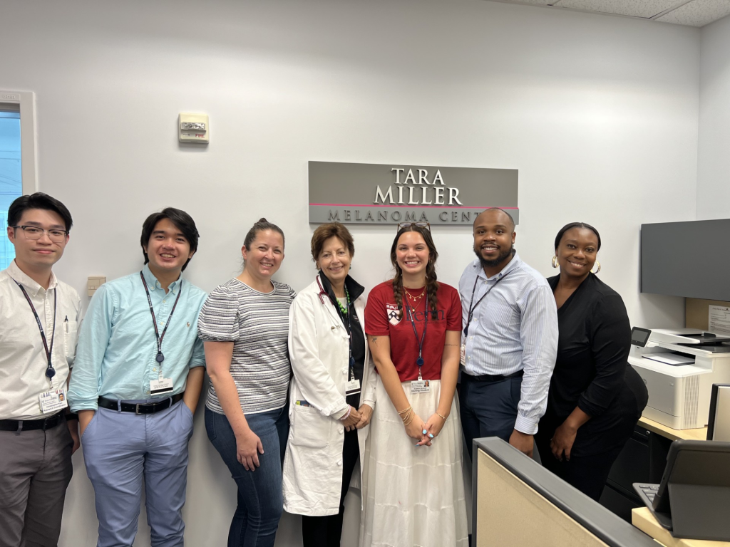 A group stands smiling in front of a "TARA MILLER MELANOMA CENTER" sign in an office setting.