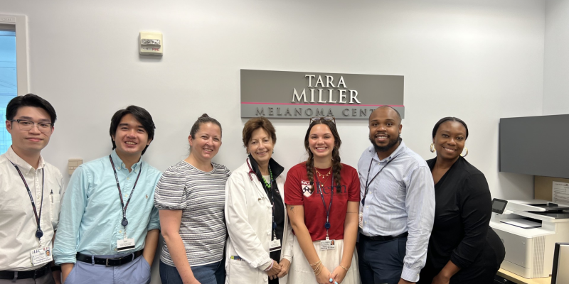 A group stands smiling in front of a "TARA MILLER MELANOMA CENTER" sign in an office setting.
