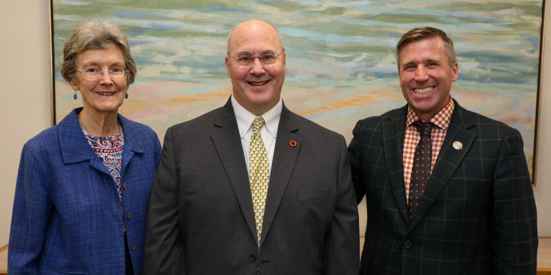 Three adults stand smiling in front of a large abstract painting, dressed in business attire.