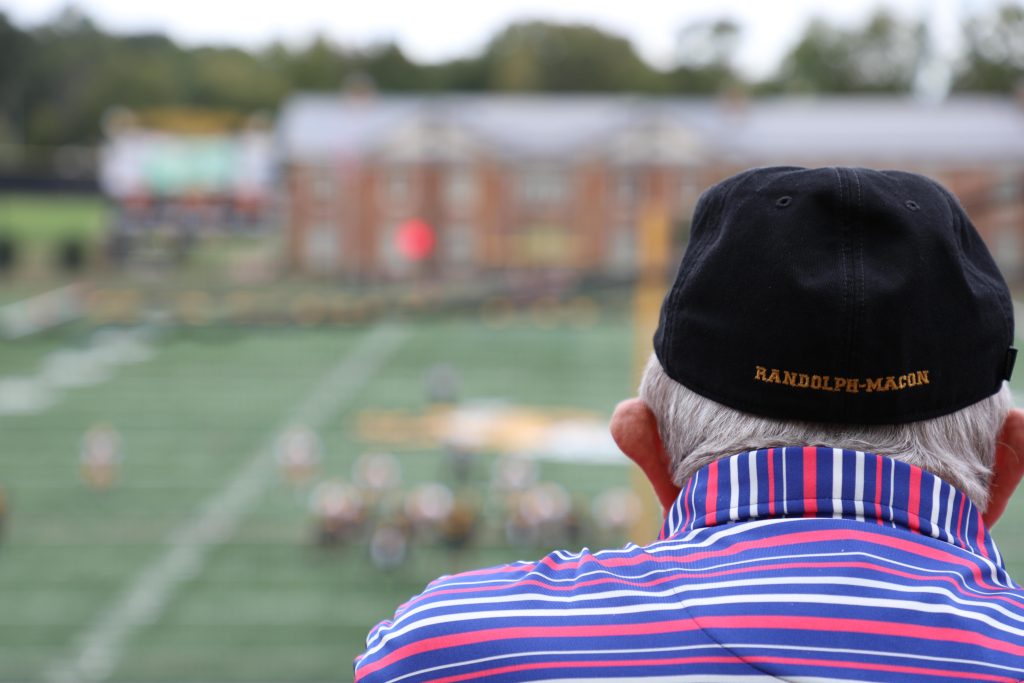 An individual wearing a “Randolph-Macon” cap observes a sports game on a field, with buildings visible in the background.