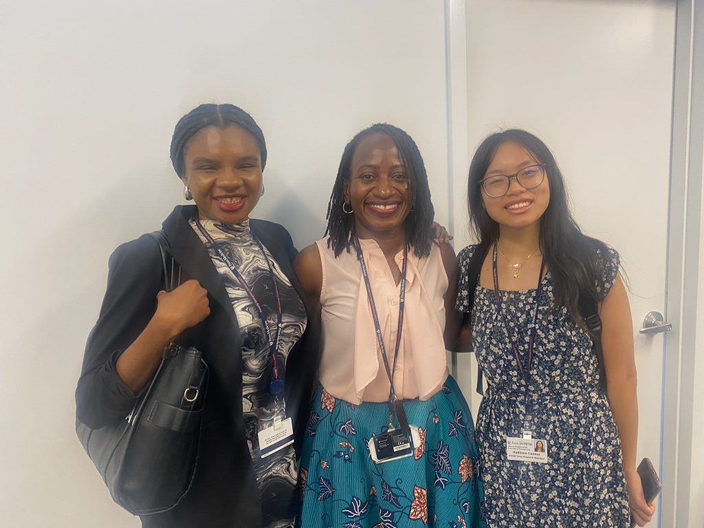 Three individuals stand together indoors, smiling at the camera and wearing name badges with professional or semi-formal attire.
