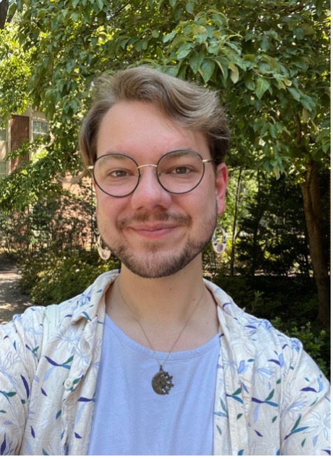 An individual smiles outdoors, wearing glasses and patterned clothing, with green trees and bushes in the background.