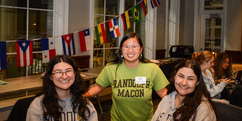 Three students pose indoors, with international flags displayed behind them. One wears a "Randolph Macon" t-shirt.
