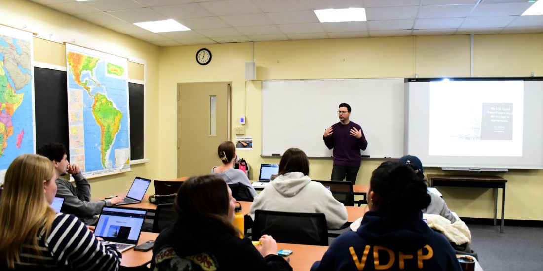 An instructor presents to a classroom of students seated at desks with laptops and notebooks.