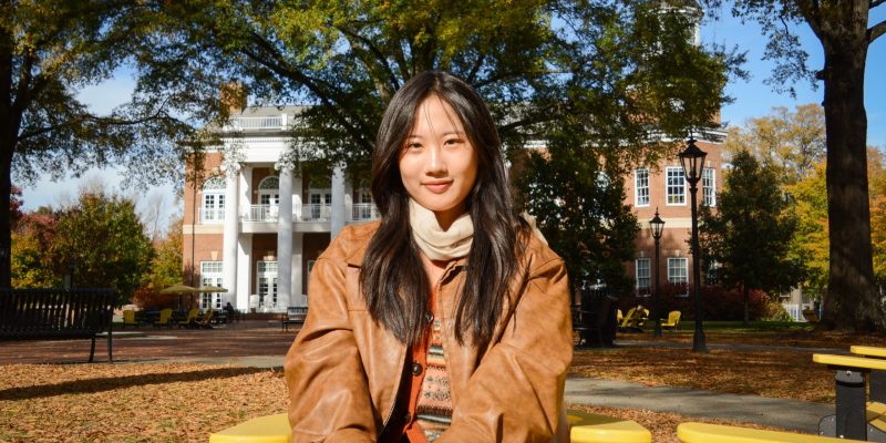 Person sits on a yellow bench in a leafy outdoor area, with a brick building and white columns in the background.