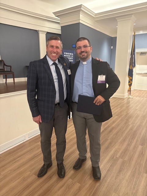 Two people stand side by side, smiling at the camera in a formal indoor setting with wood flooring and flags in the background.