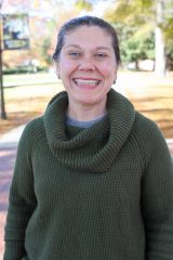 Victoria Navarra smiles outdoors on a sunny day, standing on a tree-lined path with autumn leaves in the background.