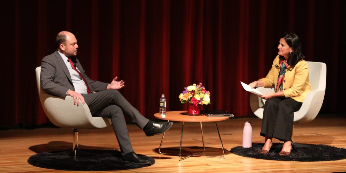 Two people sit in armchairs on stage, conversing across a small table with items. A red curtain serves as the backdrop.