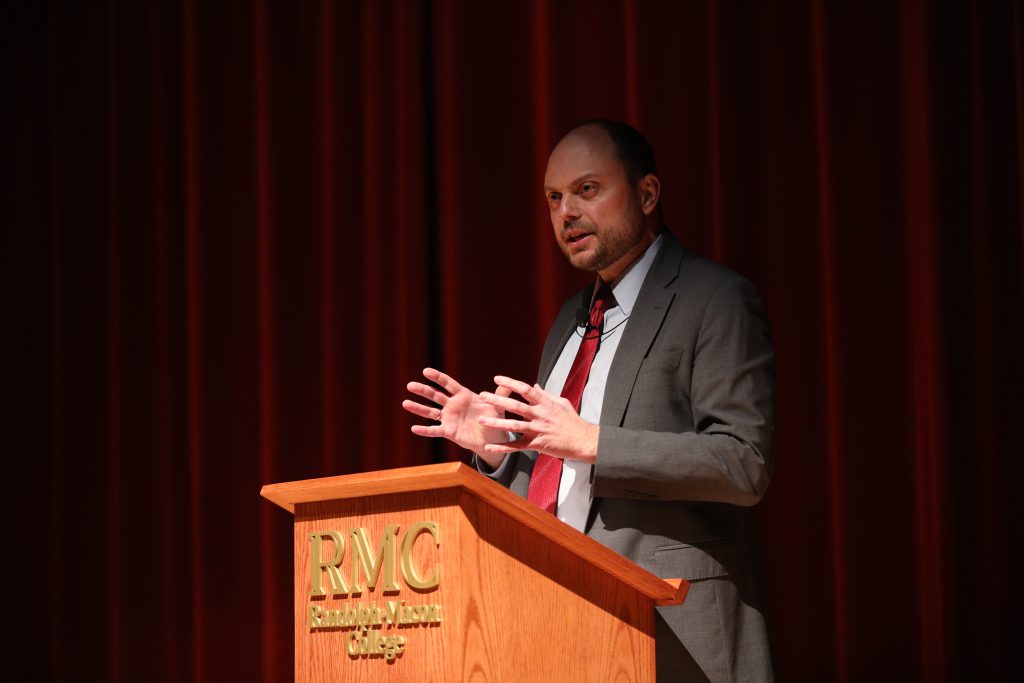 A person speaks at a podium labeled "RMC" in front of red curtains.