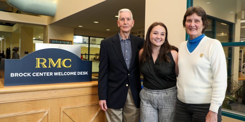 Three people stand smiling next to an RMC Brock Center Welcome Desk sign inside a building with glass windows.