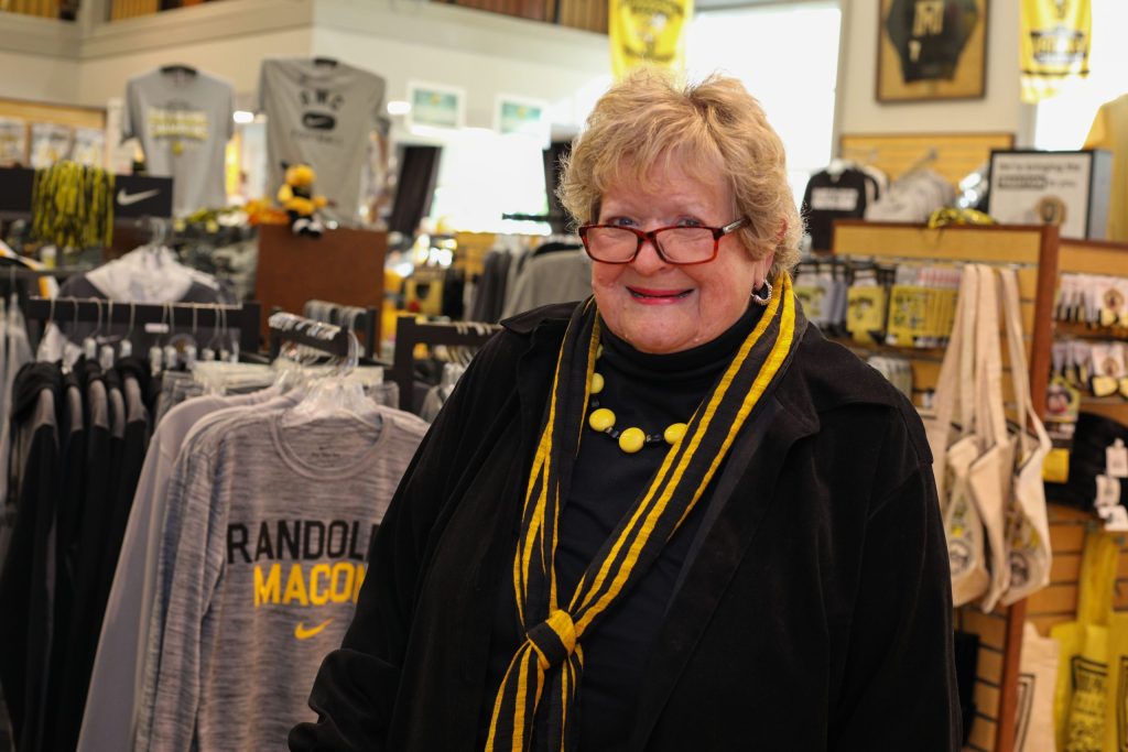 A person stands smiling in a college merchandise store with "Randolph-Macon" apparel displayed around them.