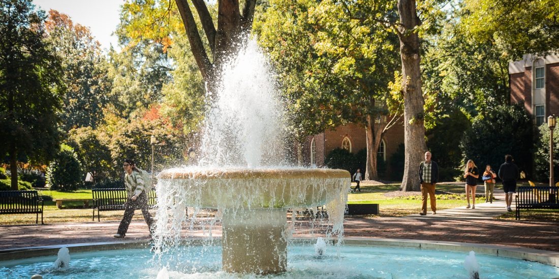 The fountain flows in the center of campus, with trees, benches, and people walking or sitting nearby.