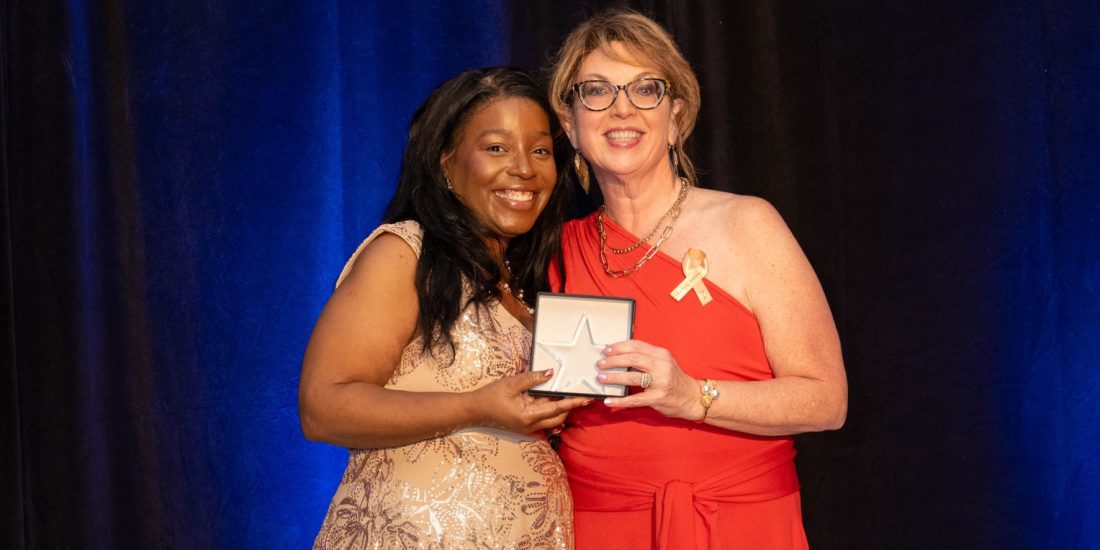 Two people stand together on stage; one holds an award. A dark curtain and blue lighting are visible in the background.