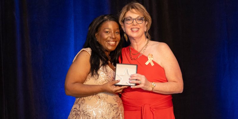 Two people stand together on stage; one holds an award. A dark curtain and blue lighting are visible in the background.