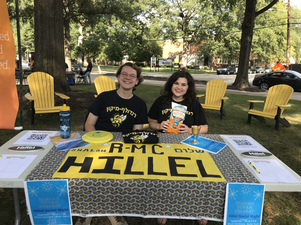 Two people behind a table with a Hillel sign offer snacks, water, and chaplain services info outdoors.
