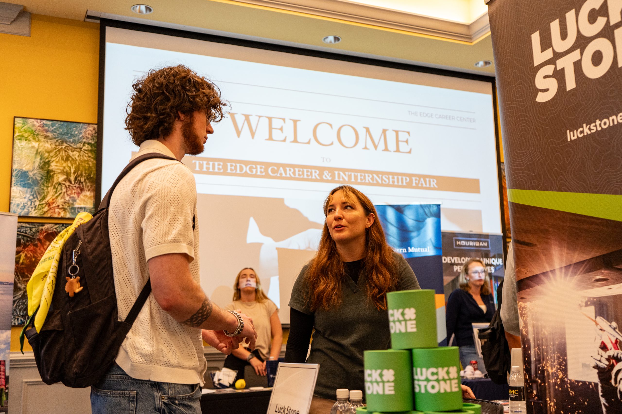 Two people converse at a booth during an Internship Fair, with "Welcome" shown on a large screen in the background.