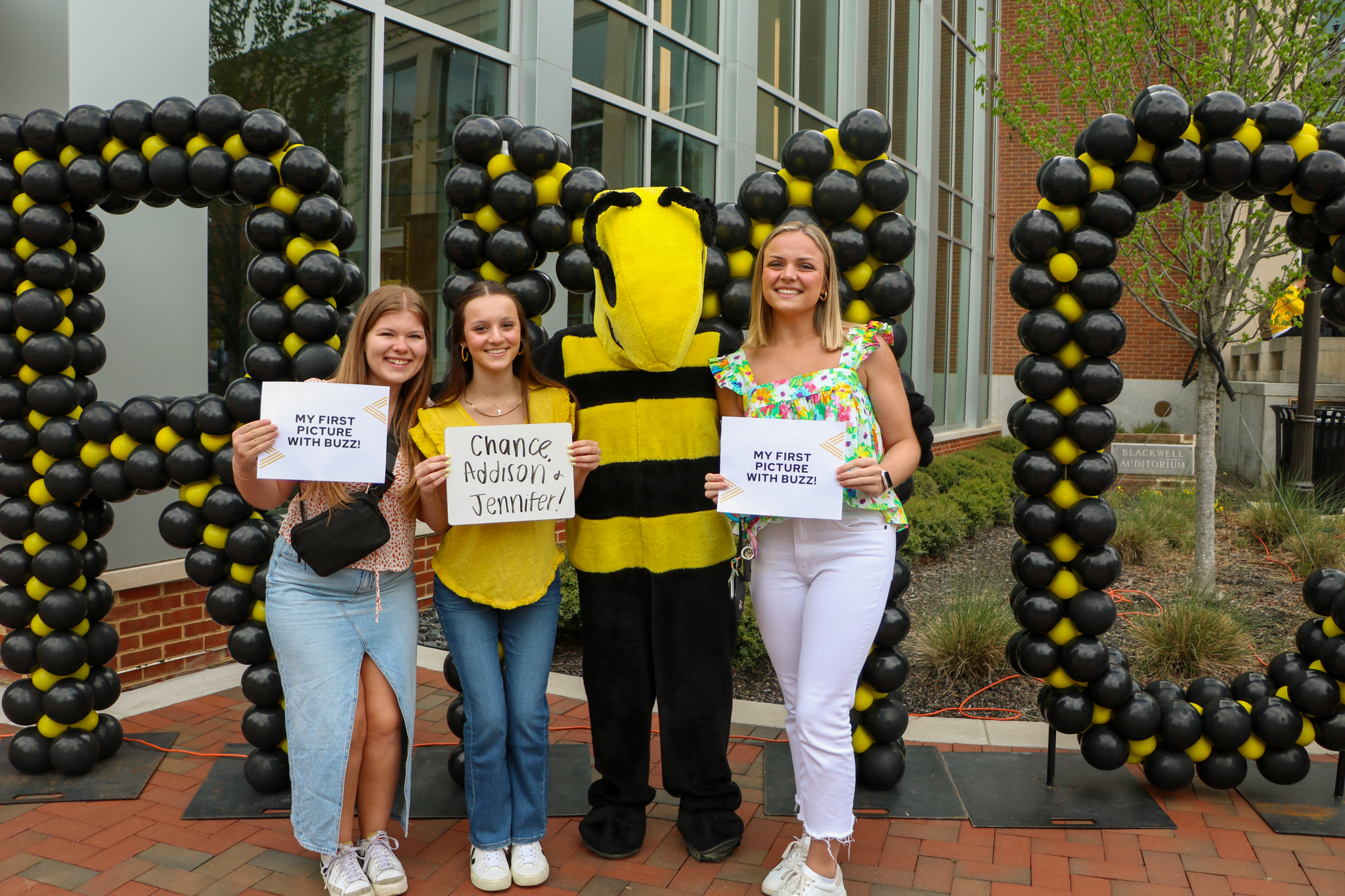 Group poses with a mascot and balloon decorations; two hold “MY FIRST PIC/SELFIE WITH BUZZ” signs, one holds an info sign.