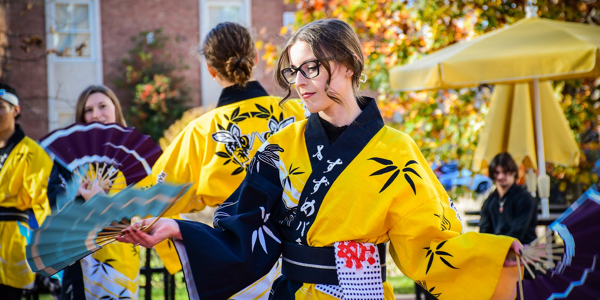 A group performs a traditional fan dance outdoors amid autumn trees and a yellow umbrella.