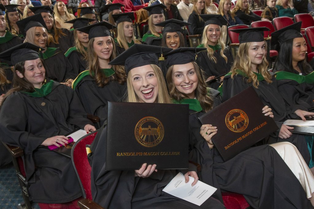 Graduates seated in an auditorium hold Randolph-Macon College diplomas and smile at the camera.
