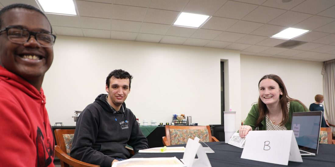 Two Mechanicsville High School students sit at a table with an RMC Education student, each with name tags, smiling during a lesson.