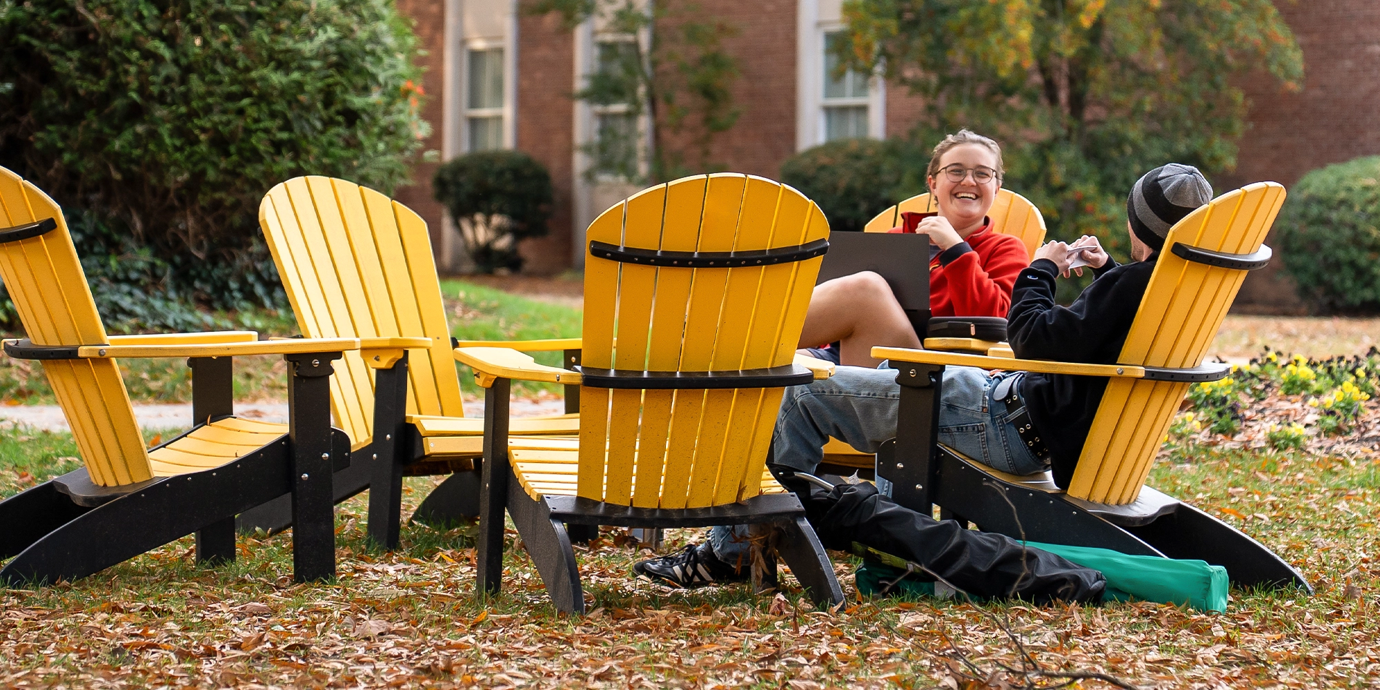 Two people converse on yellow outdoor chairs amid fallen leaves, with a building and greenery in the background.