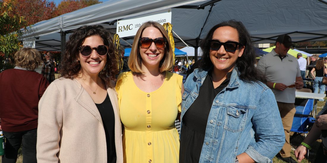 Three people smile outdoors at an event, standing in front of a tent and a crowd.