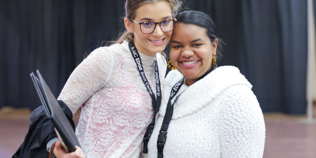 Two students wearing EDGE lanyards stand indoors, smiling at the camera. One holds folders; black curtains are in the background.