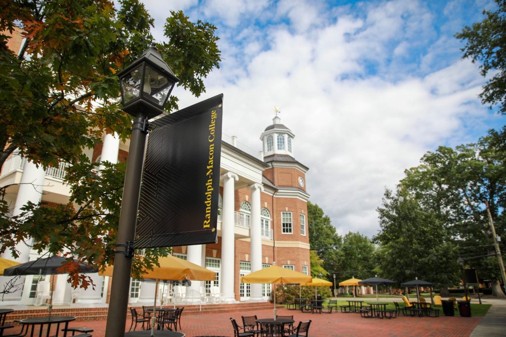 A Randolph-Macon College sign is near outdoor tables and umbrellas beside a red-brick building under a partly cloudy sky.