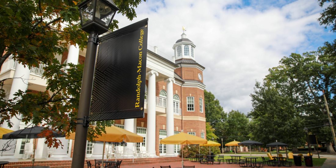 A Randolph-Macon College sign is near outdoor tables and umbrellas beside a red-brick building under a partly cloudy sky.