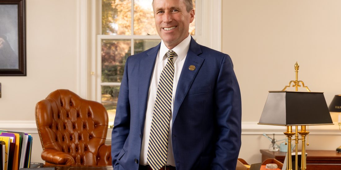 Dr. Hill smiling while standing in an office with a large wooden desk, brown leather chair, and various office items